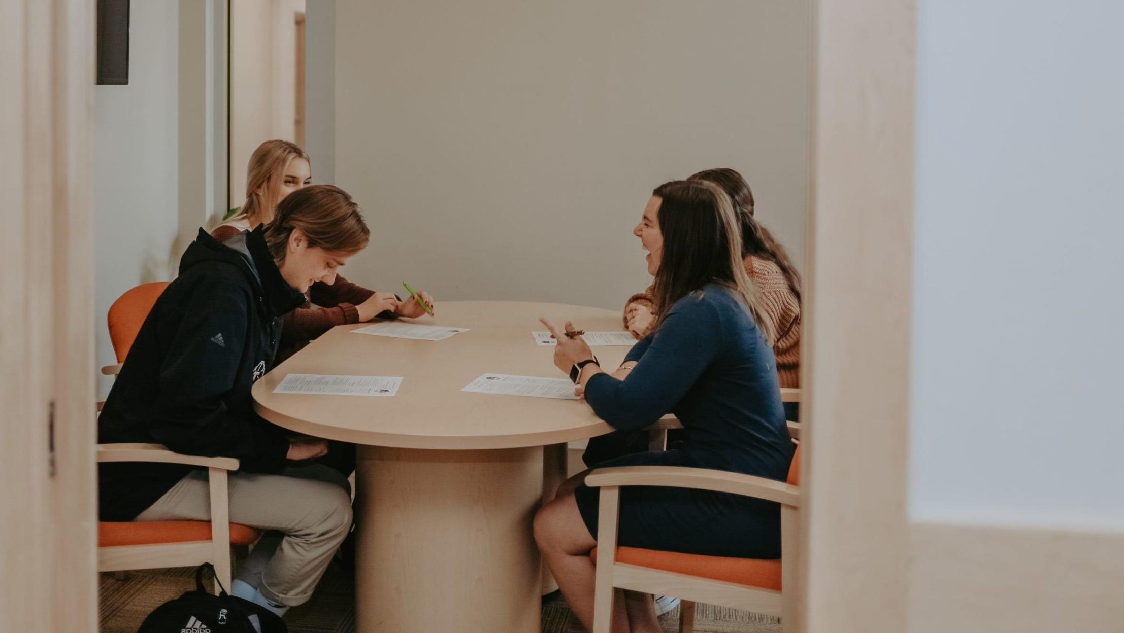 Three students sit around at table with a staff member; all four are looking at pieces of paper on the table